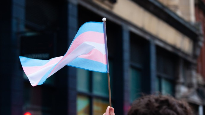 A handheld trans pride flag being waved by a hand that's just out of shot.