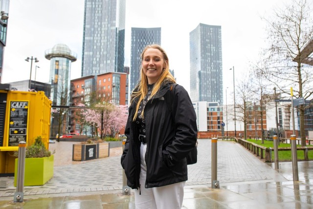 A woman student standing in the streets with the Manchester cityscape in the background. She is similing and it is raining slightly.