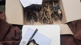 Photograph of a fire-making kit: a box containing wooden sticks tied together, some written instructions on paper and some matches laid out on some brown cloth.