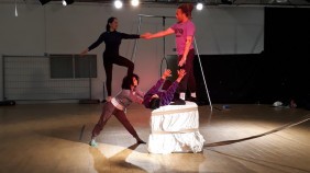 Five performers, one out of frame, stand on and to the side of an empty wooden plinth creating a carefully balanced configuration of bodies. Situated in a workshop theatre rehearsal space and lit by white stripe lights and a pink neon spotlight.