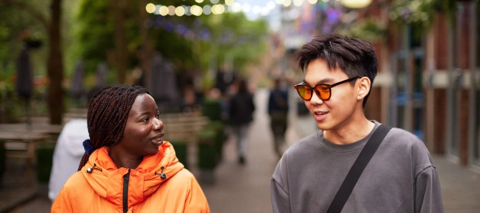 Two students walking along a road in Manchester city centre, with passersby and greenery in the background.