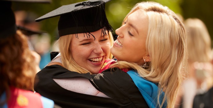 Two students in mortar boards and gowns smiling and hugging at a graduation ceremony