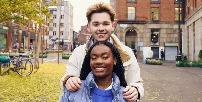 Two students smiling in the city centre, he's stood behind her with hands on her shoulder