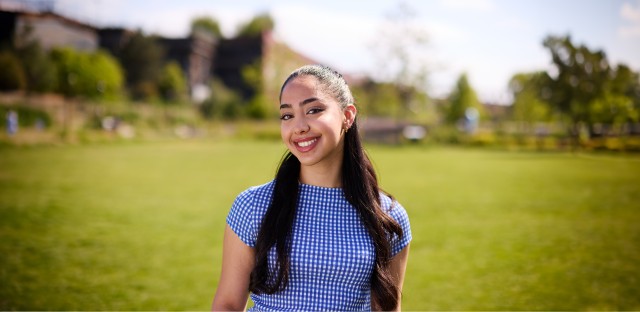A student stood in a green, grassy field smiling. There are trees and buildings in the distance behind her.